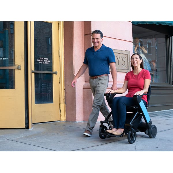 Femme assise dans un fauteuil roulant électrique WHILL Model F en centre-ville, accompagnée d’un homme marchant à ses côtés et tenant sa main.