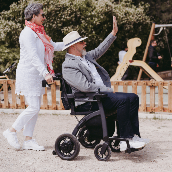 Un homme profite d’une promenade assis dans un fauteuil roulant électrique Rollz Motion Electric, accompagné et poussé par une femme, dans un parc ensoleillé.